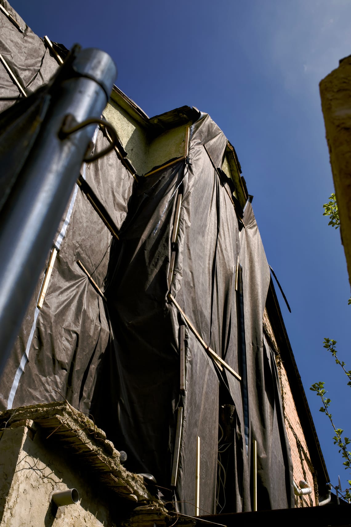 Formerly adjacent facade of a building covered with black foil and wooden rods. The demolished building leaves an open space and blue sky can be seen.